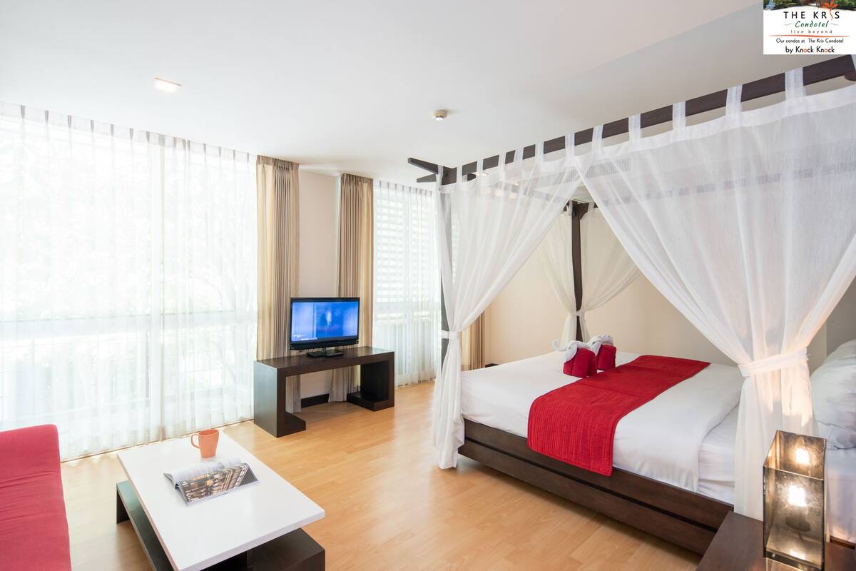 Una habitación de hotel moderna con una cama grande, televisión, una mesita de centro y vistas a través de cortinas translúcidas.
