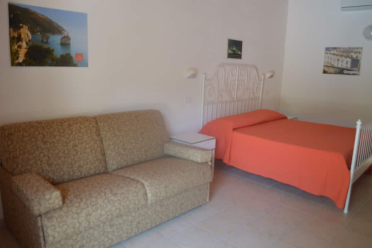 A bedroom with a patterned sofa, a single bed with an orange bedspread, a white headboard, and a small bedside table.