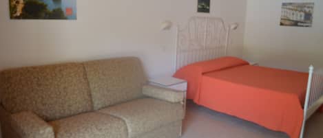 A bedroom with a patterned sofa, a single bed with an orange bedspread, a white headboard, and a small bedside table.