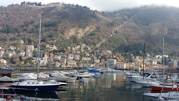 Marina com barcos atracados, um morro com edifícios e montanhas ao fundo.