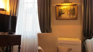 Uma sala de estar com televisão sobre uma mesa de madeira, uma poltrona bege, um radiador e um quadro na parede.
