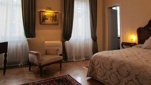 Quarto de hotel com cama, uma cadeira, escrivaninha e um quadro na parede.