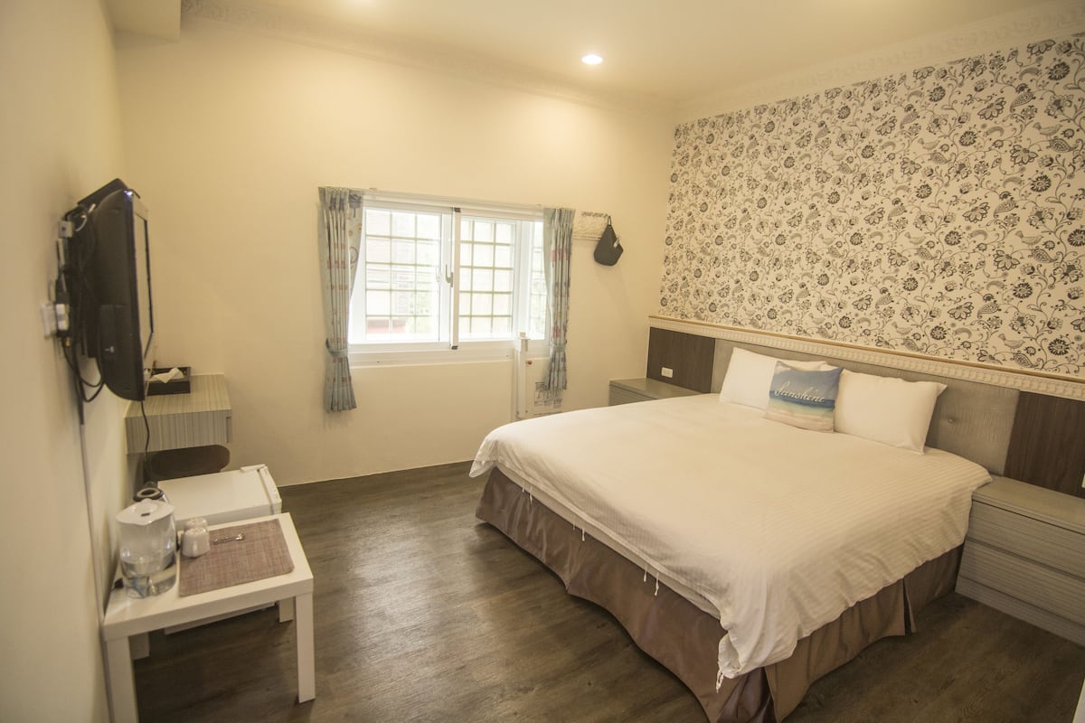 Un dormitorio con una cama grande, una mesita de noche y cortinas en la ventana. La pared presenta un diseño decorativo.