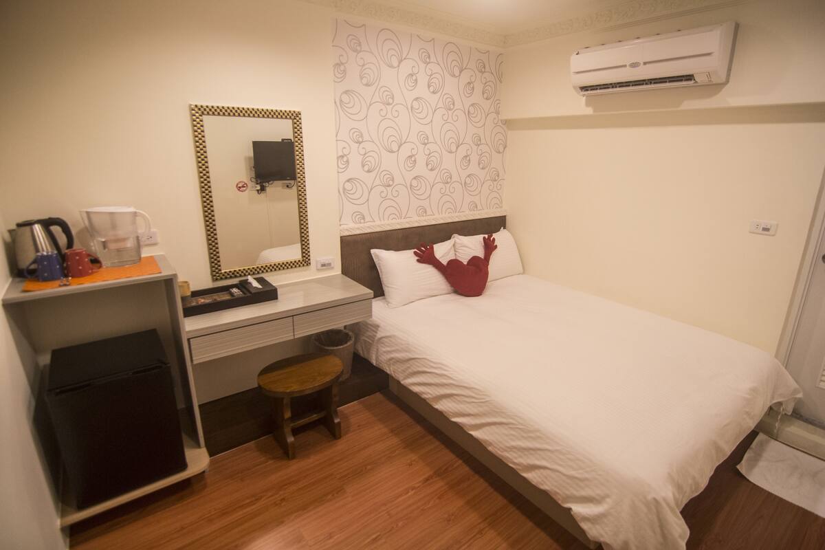 Una habitación de hotel con cama, escritorio con silla, un taburete pequeño, un espejo y un equipo de aire acondicionado.