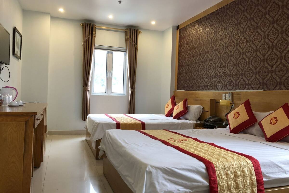 A hotel room with two beds, a wooden desk, a window with curtains, and a patterned wall.