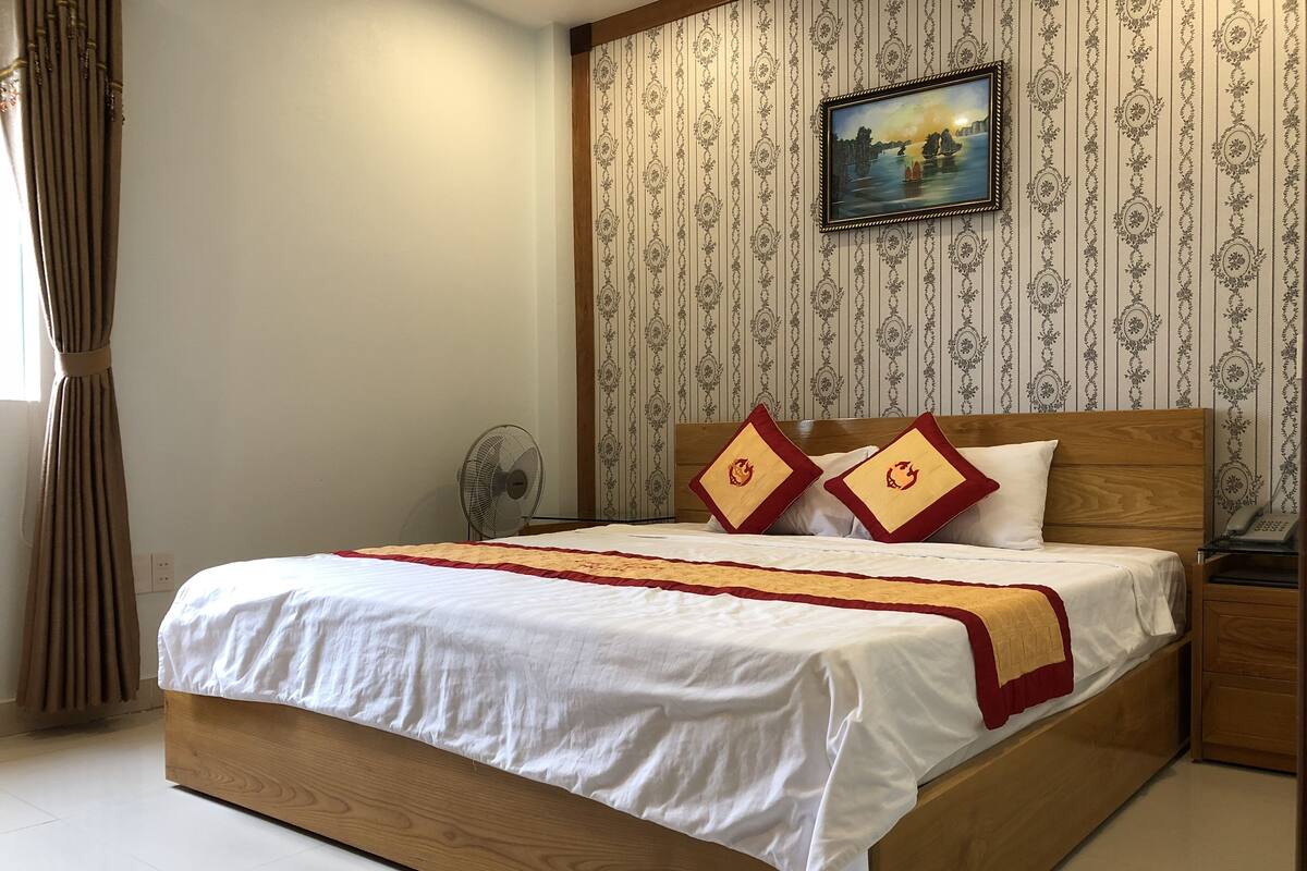A neatly made bed with a wooden frame, a white bedspread, and two red pillows with a lion emblem. A fan is on the floor beside the bed, and a painting hangs on the wall above it.