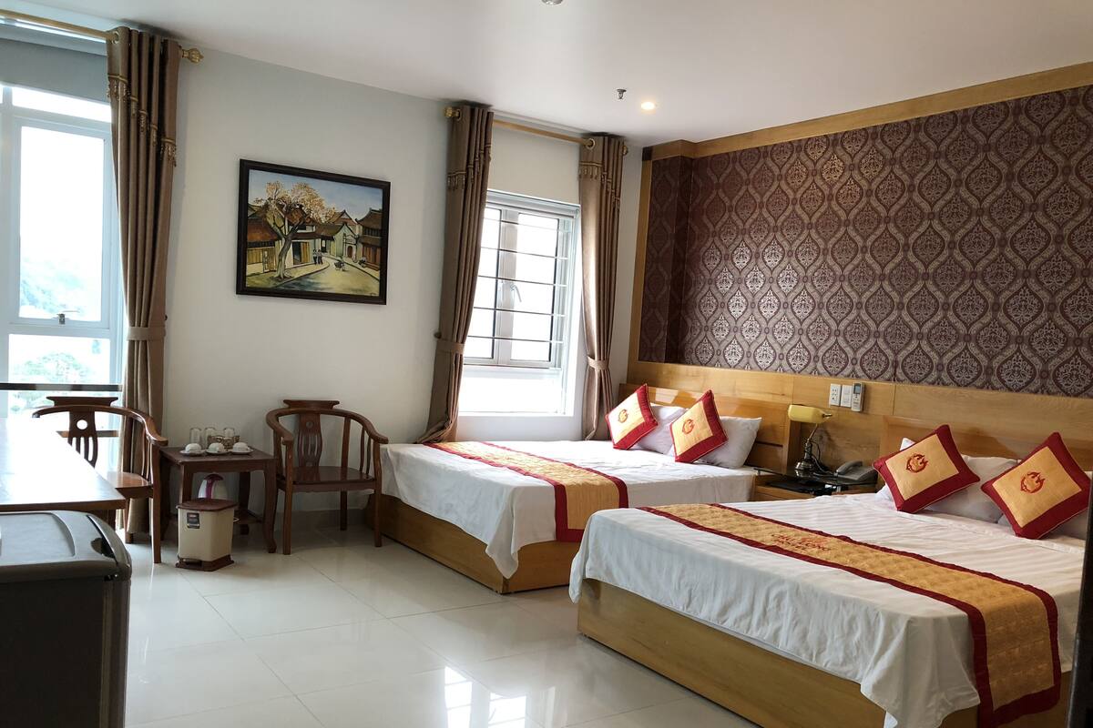 A hotel room with two beds, a desk, chairs, a trash bin, and a window with curtains.