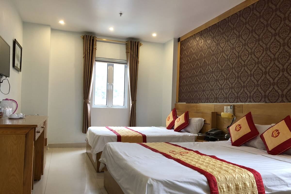 A hotel room with two beds, a wooden desk, a window with curtains, and a patterned wall.
