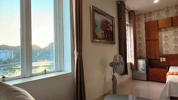 Una habitación con un sofá, una mesa con tazas de té, un ventilador, un refrigerador y una ventana con vistas a edificios y colinas.