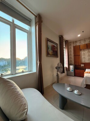 A room with a sofa, a table with teacups, a fan, a refrigerator, and a window with a view of buildings and hills. - Chu Long Hotel & Spa (Hai Phong)