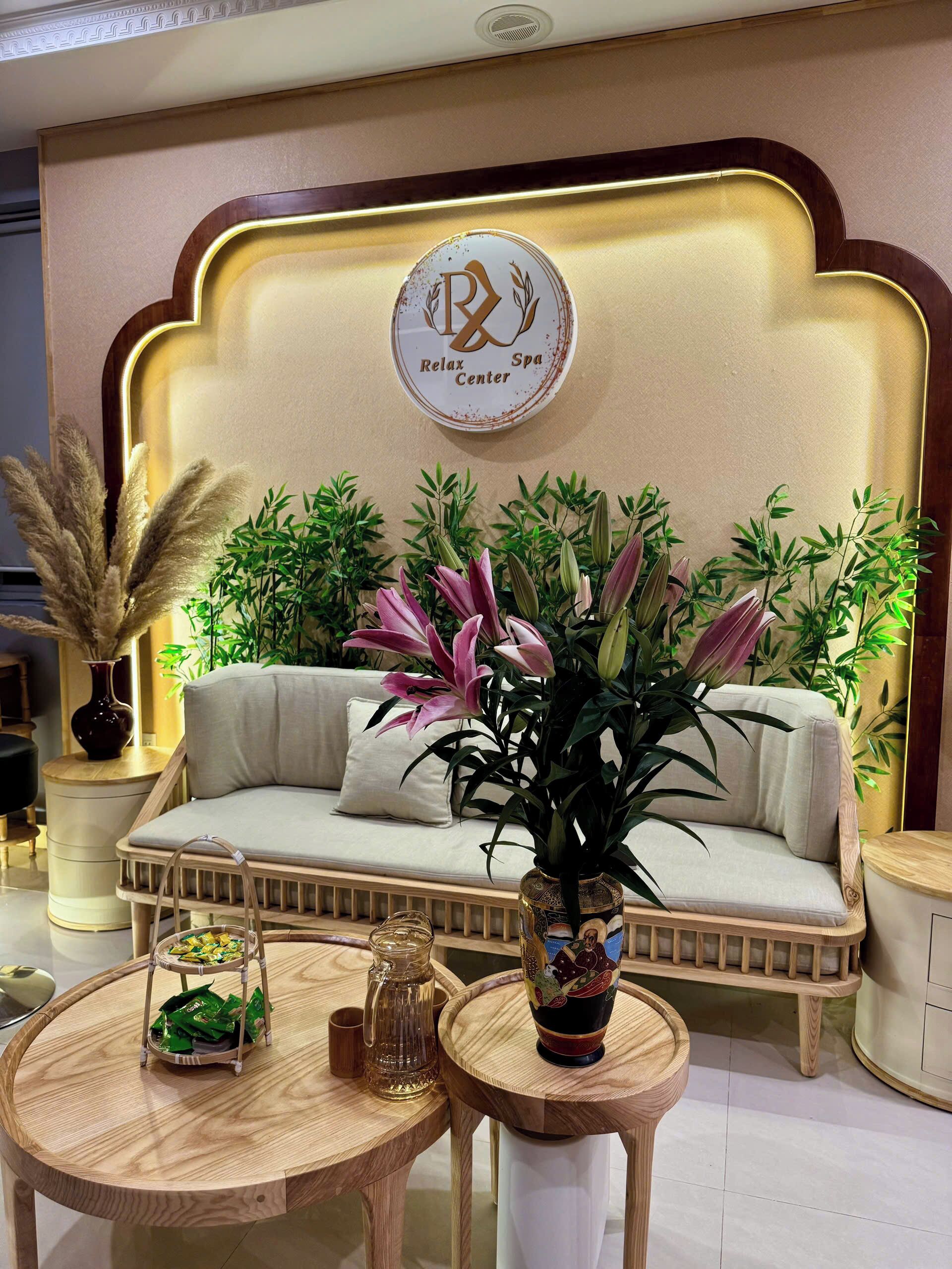 A lounge area with a sofa, decorative plants, a wall-mounted sign, and a small table with a pitcher and a vase.