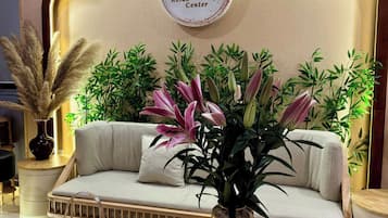 A lounge area with a sofa, decorative plants, a wall-mounted sign, and a small table with a pitcher and a vase.