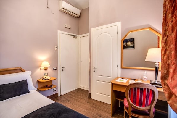 A hotel room with a bed, bedside table with a lamp, a desk with a chair, a mirror, and a door. - San Valentino (Rome)