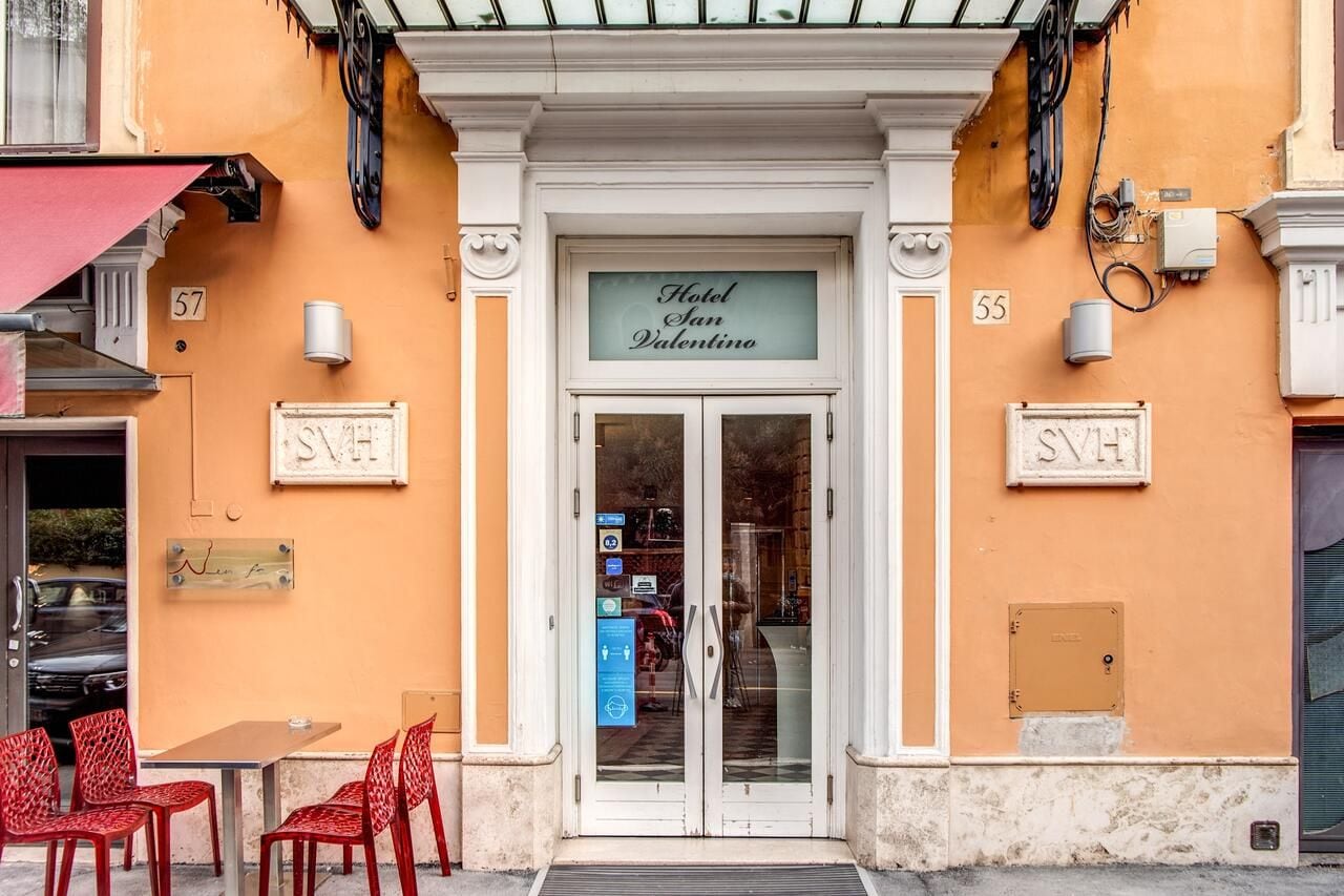 Entrada do Hotel San Valentino, com uma placa acima da porta, uma pequena área de estar ao ar livre com cadeiras vermelhas e o número '55' na parede.