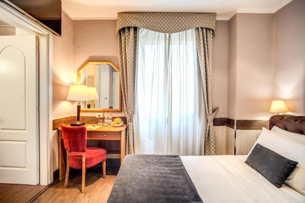 Una habitación de hotel con cama, escritorio, silla, espejo y ventana con cortinas.