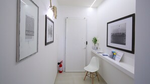 A hallway with framed pictures, a white door, a white chair, and a shelf with books and a plant.