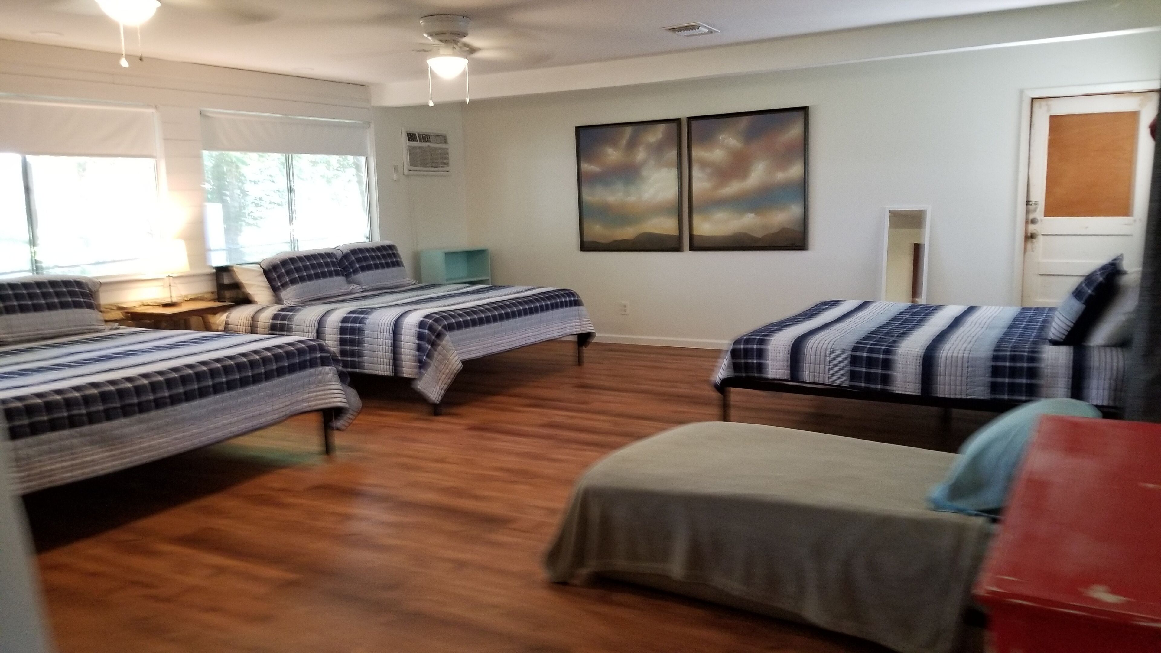A room with three beds, wooden flooring, a desk with a lamp, and two paintings on the wall.
