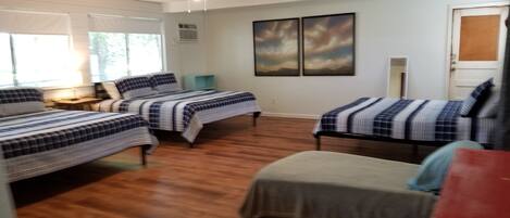 A room with three beds, wooden flooring, a desk with a lamp, and two paintings on the wall.