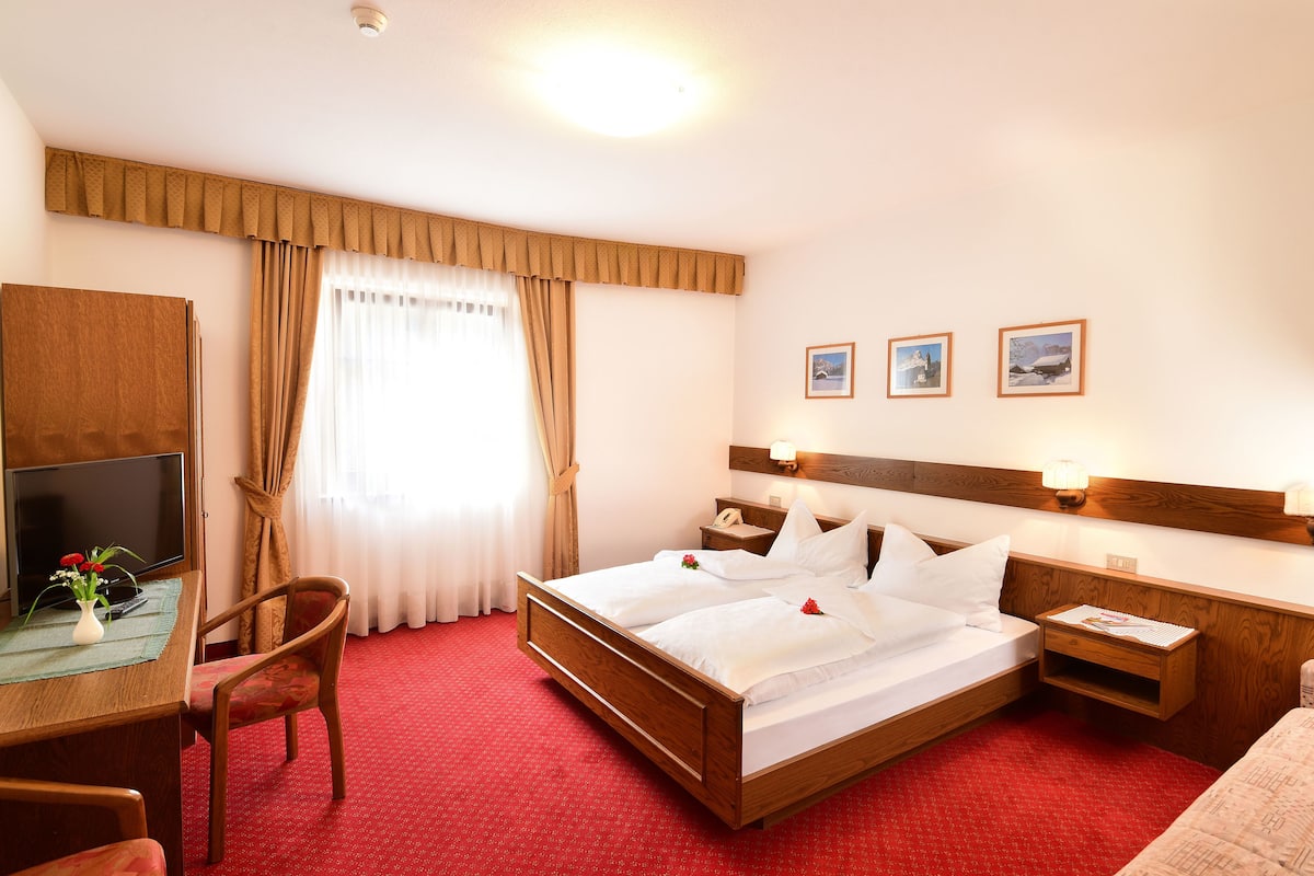 A hotel room with a wooden bed, a desk with a TV, a chair, and a red carpet.