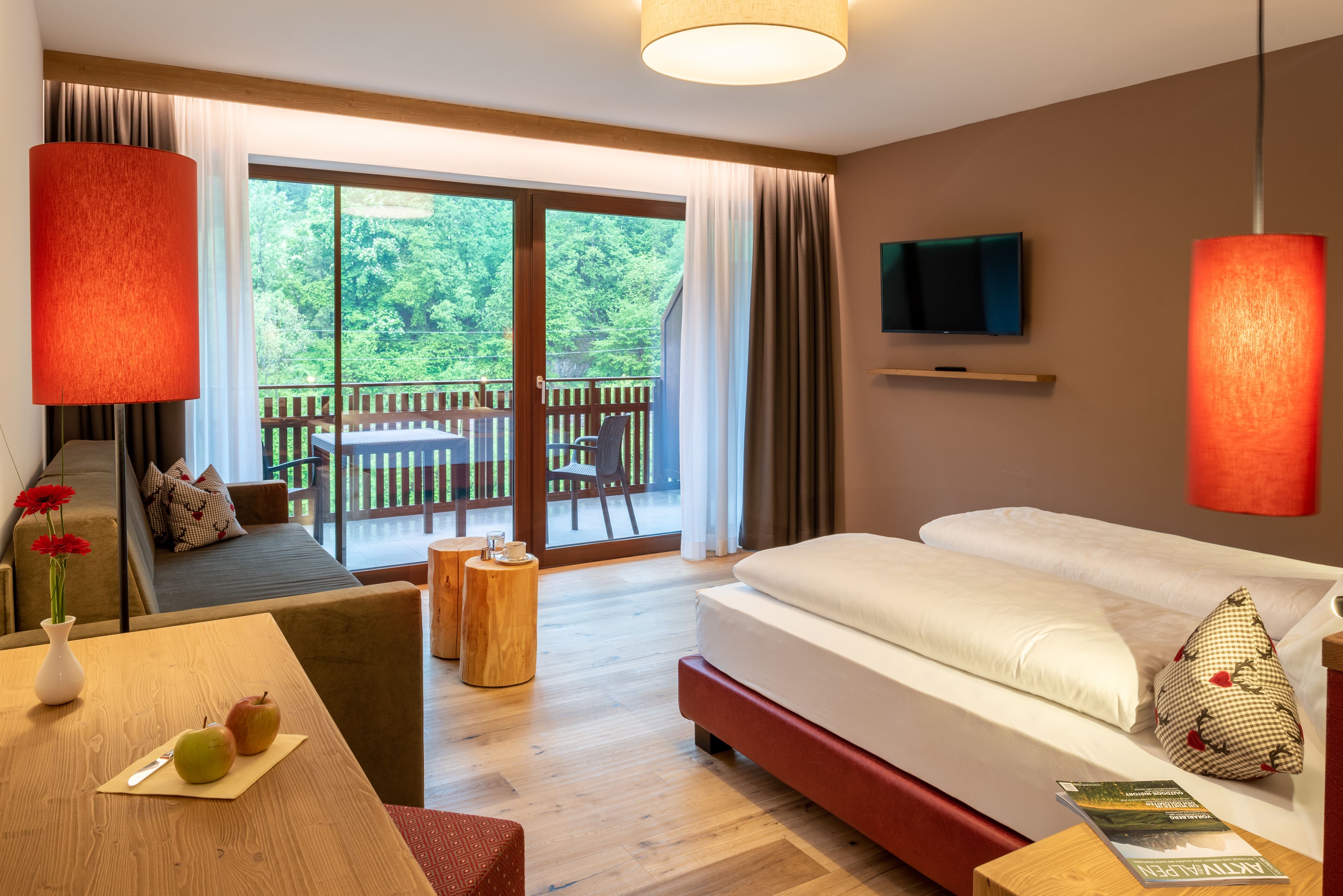 A modern hotel room with a large sliding glass door to a balcony, a sofa, a bed, a wooden side table, and a TV mounted on the wall.