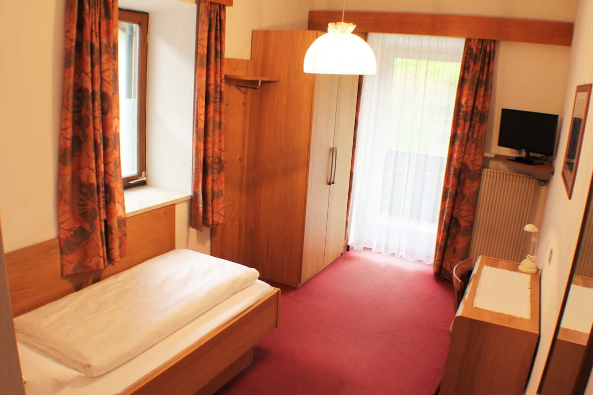 A hotel room with a single bed, wooden wardrobe, a desk with a mirror, a television, and a window with curtains.