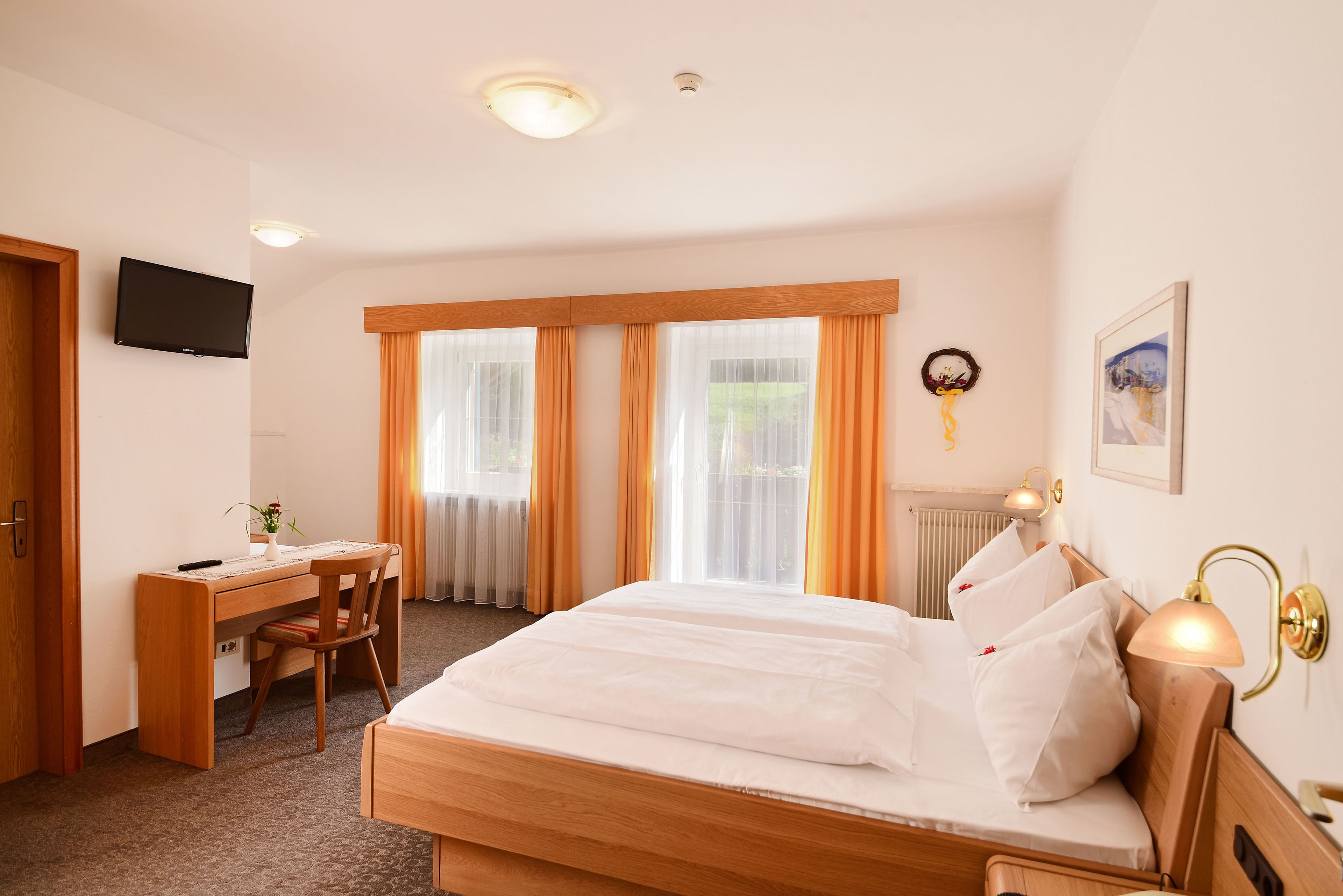 A hotel room with a wooden bed, a desk with a chair, a television on the wall, and a window with curtains.