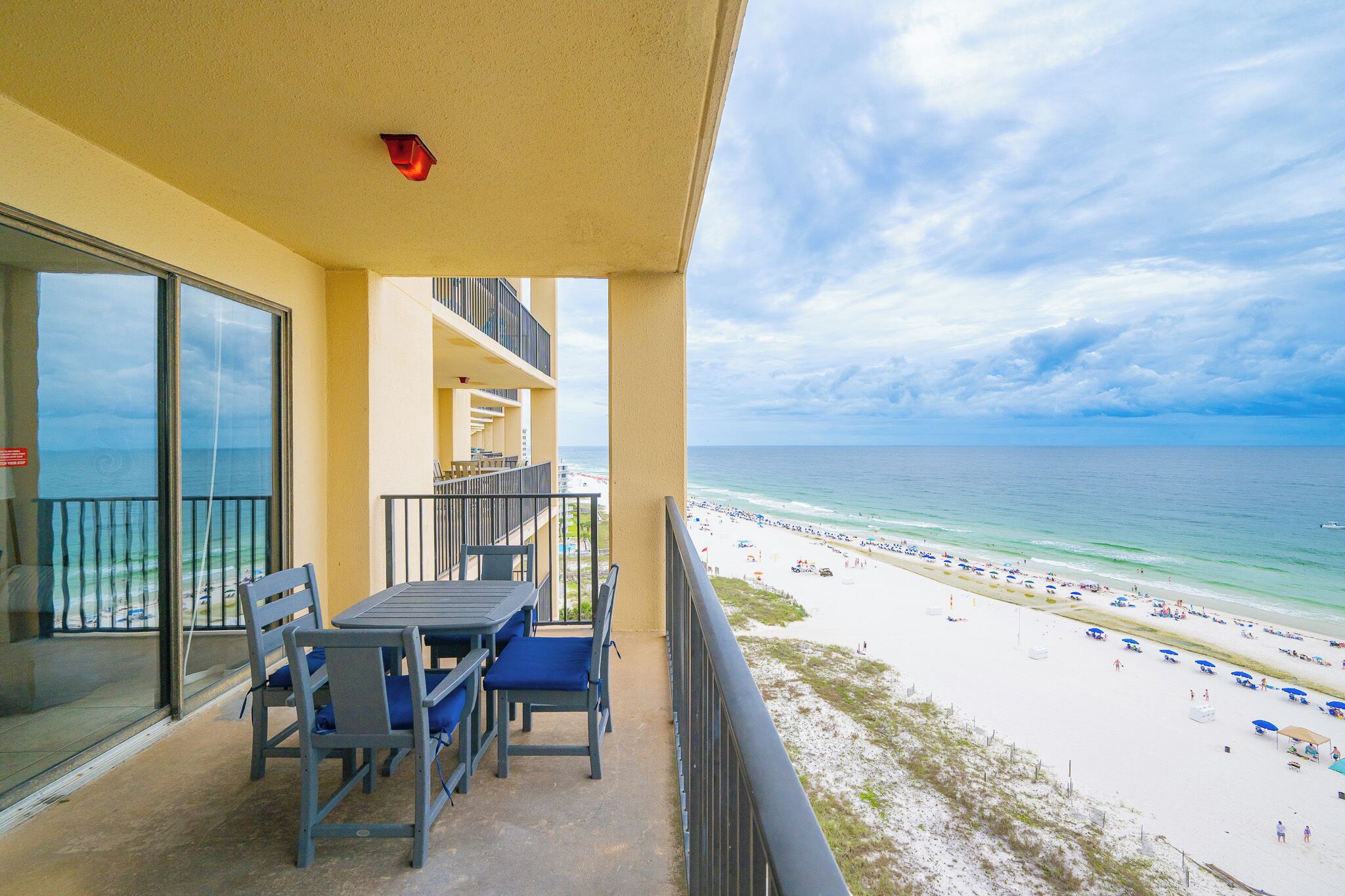 A balcony with a table and chairs, overlooking a beach with umbrellas and people.