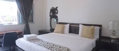 A hotel room with a large bed, a desk, a chair, a nightstand, a lamp, and a window with curtains.