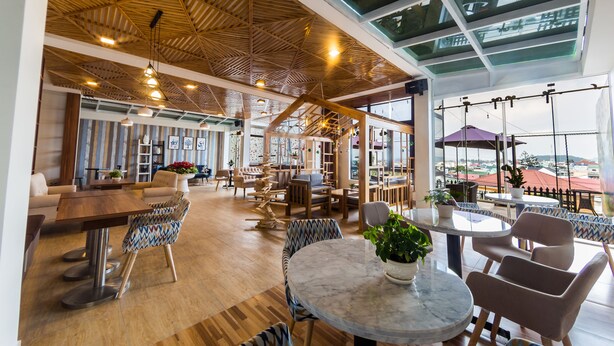 Un vaste espace intérieur doté d’un plancher en bois, de plusieurs coins salon comprenant des tables et des chaises, et de grandes fenêtres offrant une vue sur la ville.