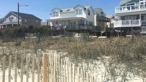 Een strandhuis met een houten terras, een zandstrand op de voorgrond en een helderblauwe lucht.