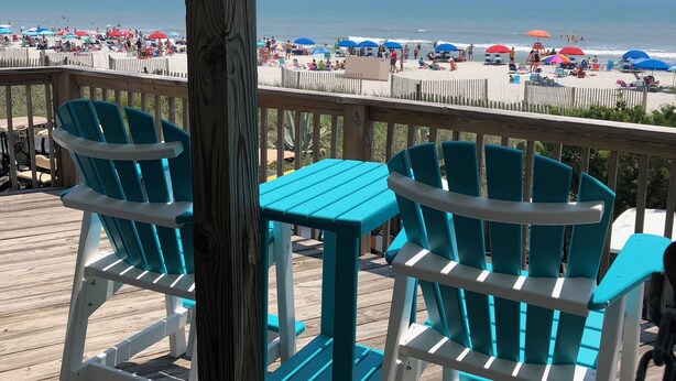 Een houten terras met turquoise Adirondack-stoelen en een kleine tafel, met uitzicht op een strand met parasols en mensen.
