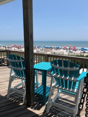 A wooden deck with turquoise Adirondack chairs and a small table, overlooking a beach with umbrellas and people.