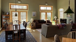 A spacious living area with a wooden ceiling, large windows, a dining table with chairs, a sofa, and a television.