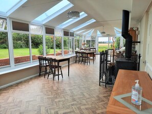 A spacious room with large windows, a wooden table, chairs, and a black stove. - Elmfield House B&B (Bedale)
