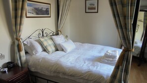 Een netjes opgemaakt bed met witte lakens, een nachtkastje met een radio en een wandklok.