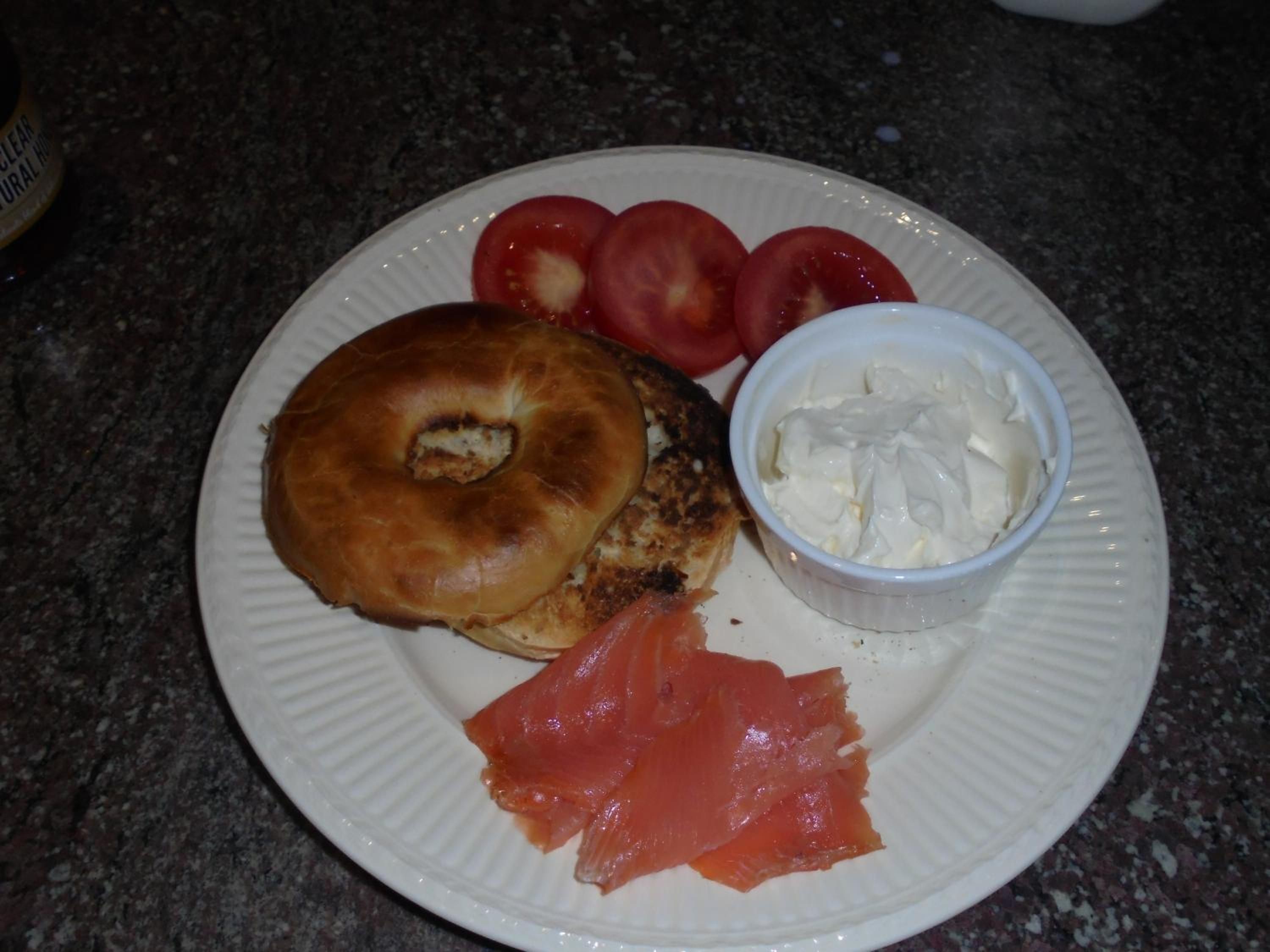 A plate with a bagel, smoked salmon, tomatoes, and a small container of cream cheese.