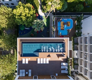 An aerial view of a modern hotel with a large rectangular swimming pool surrounded by lounge chairs and a wooden deck.