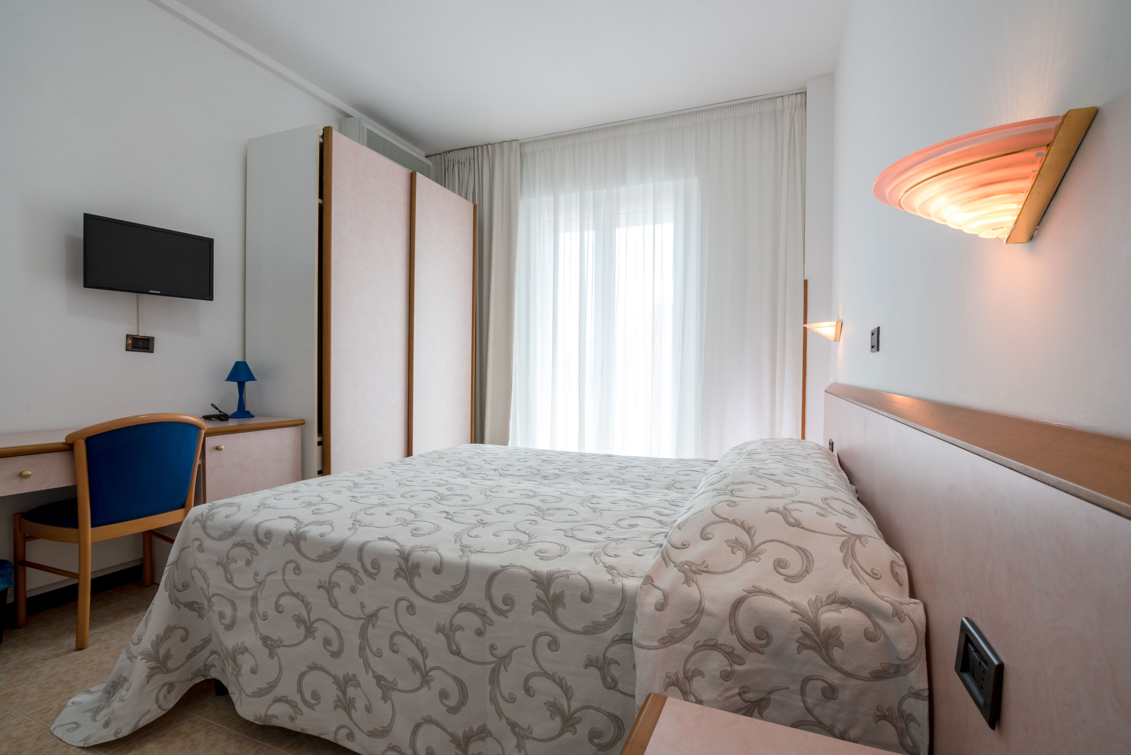 A hotel room with a bed, a desk with a chair, a television, and a window with curtains.