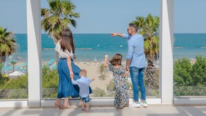 Uma família de quatro pessoas em pé perto de uma grande janela com vista para uma praia, palmeiras e um céu azul límpido.
