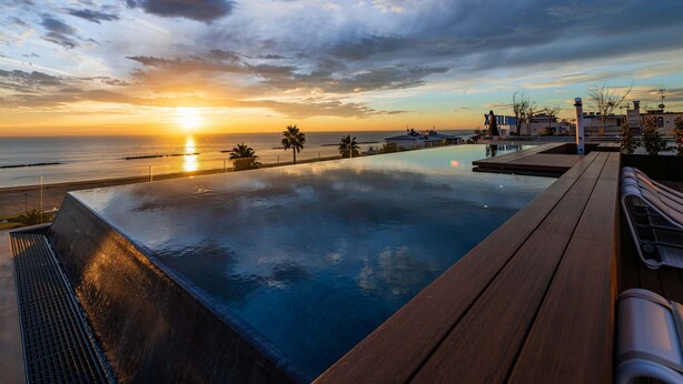 Uma piscina infinita com um deck de madeira, com vista para o pôr do sol sobre o oceano, com edifícios ao fundo.