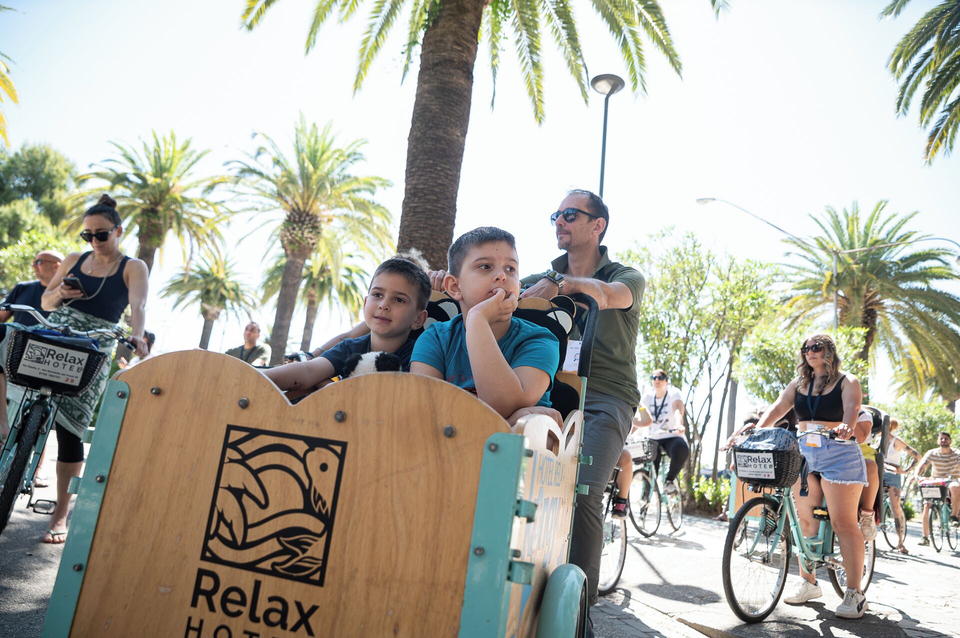 Un carretto di legno con il logo Relax Hotel, circondato da persone e biciclette in un'ambientazione esterna assolata.