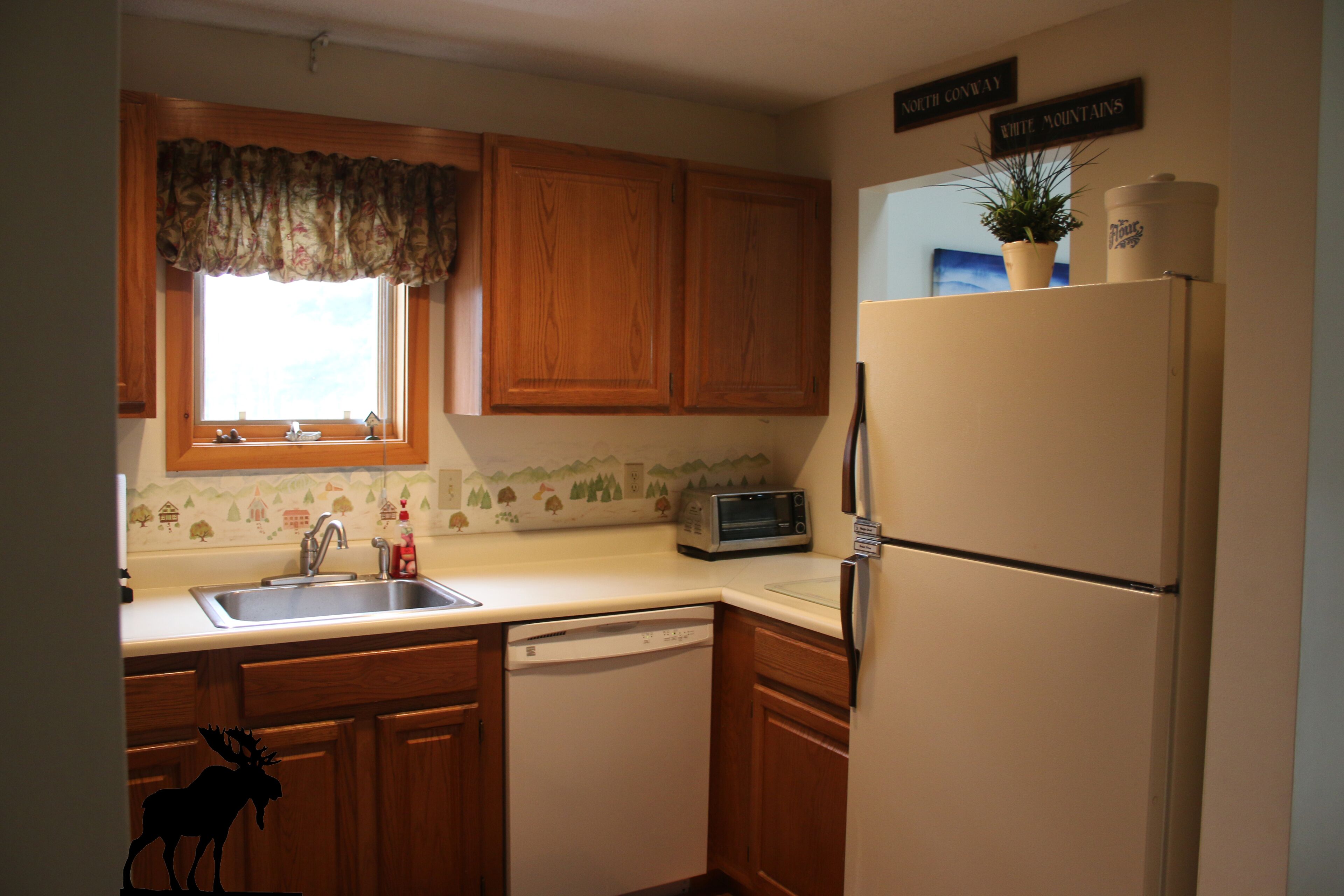 A kitchen with wooden cabinets, a dishwasher, a refrigerator, a toaster, and a window with a curtain.