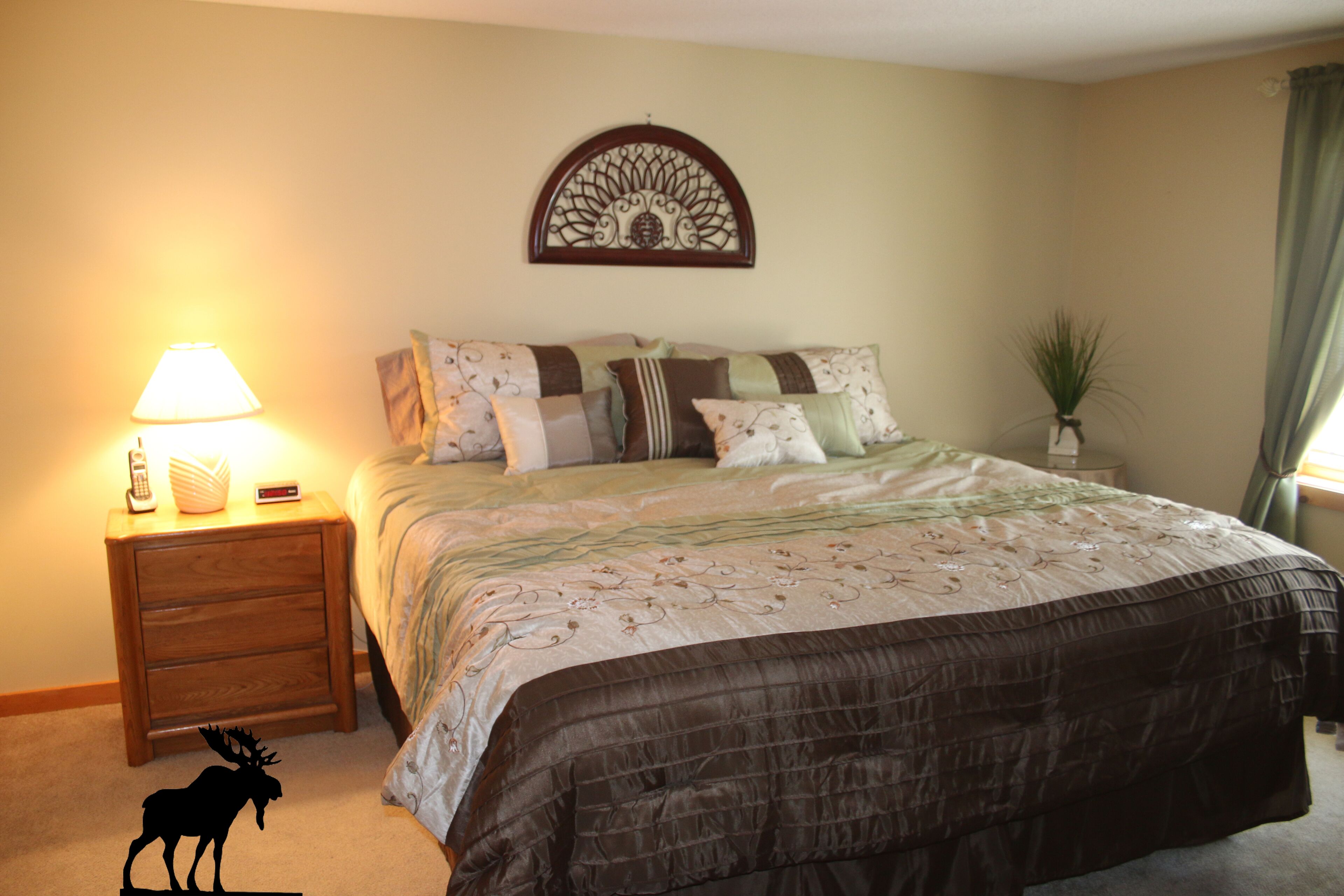A bedroom with a bed, a nightstand with a lamp, a decorative wall piece, and a small plant.