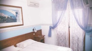Quarto de hotel com cama, mesa de cabeceira, luminária, quadro na parede e janela com cortinas translúcidas.