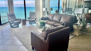 Una sala de estar moderna con una mesa de centro de vidrio, sofá de cuero y sillones, con vista a un balcón y al mar.