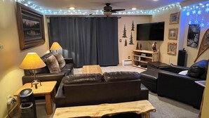 A living room with a dark leather sofa, a wooden coffee table, and a TV on a stand. There are string lights on the ceiling and a ceiling fan.