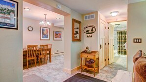 A hallway with a wooden dining table and chairs, a small wooden side table with a bowl, a welcome sign with a bear and trees, and a bathroom visible through an open door.