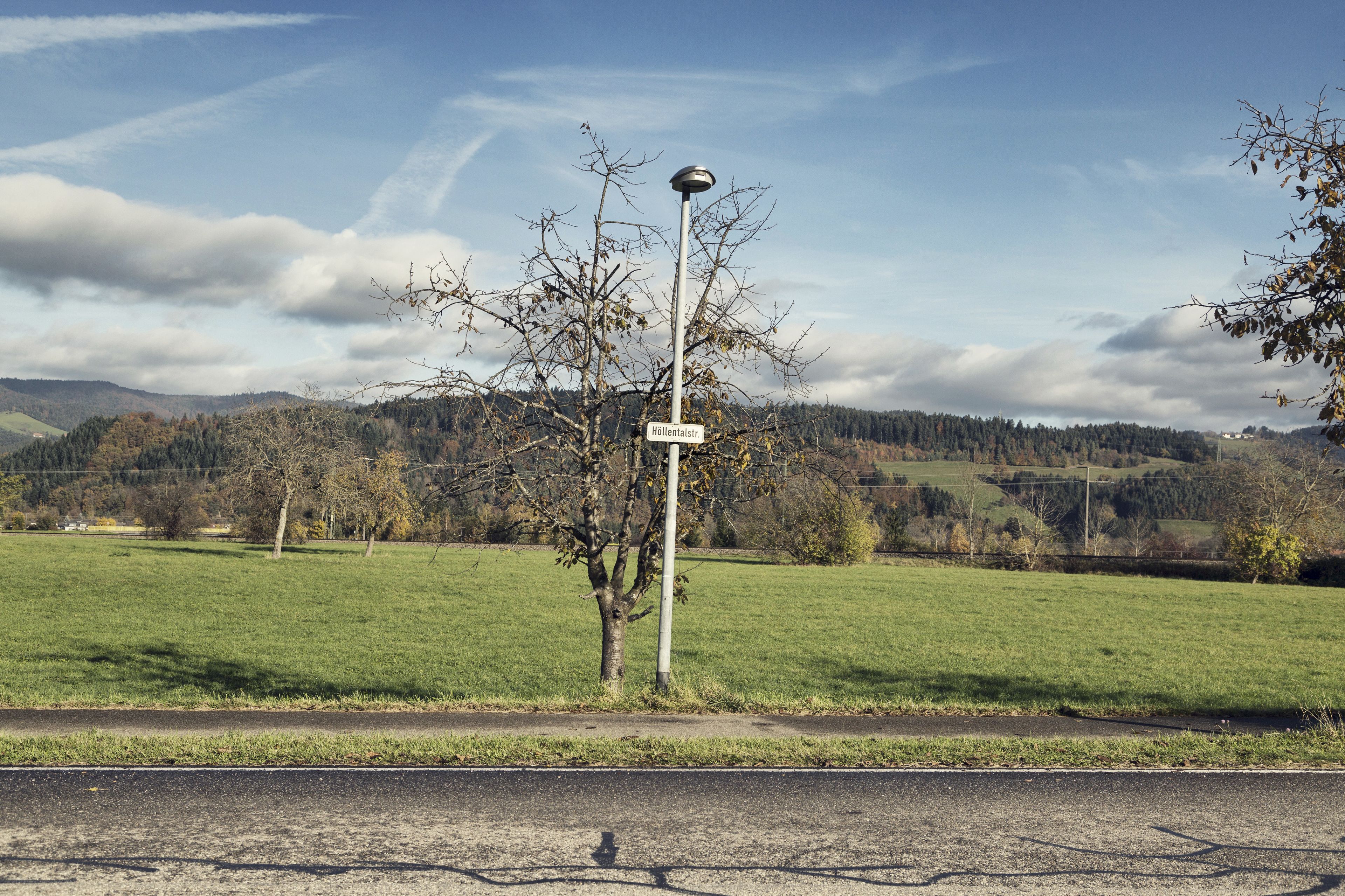 Una señal de calle que muestra 'Hollentalstr.', en un entorno rural con cielo despejado y un paisaje montañoso al fondo.