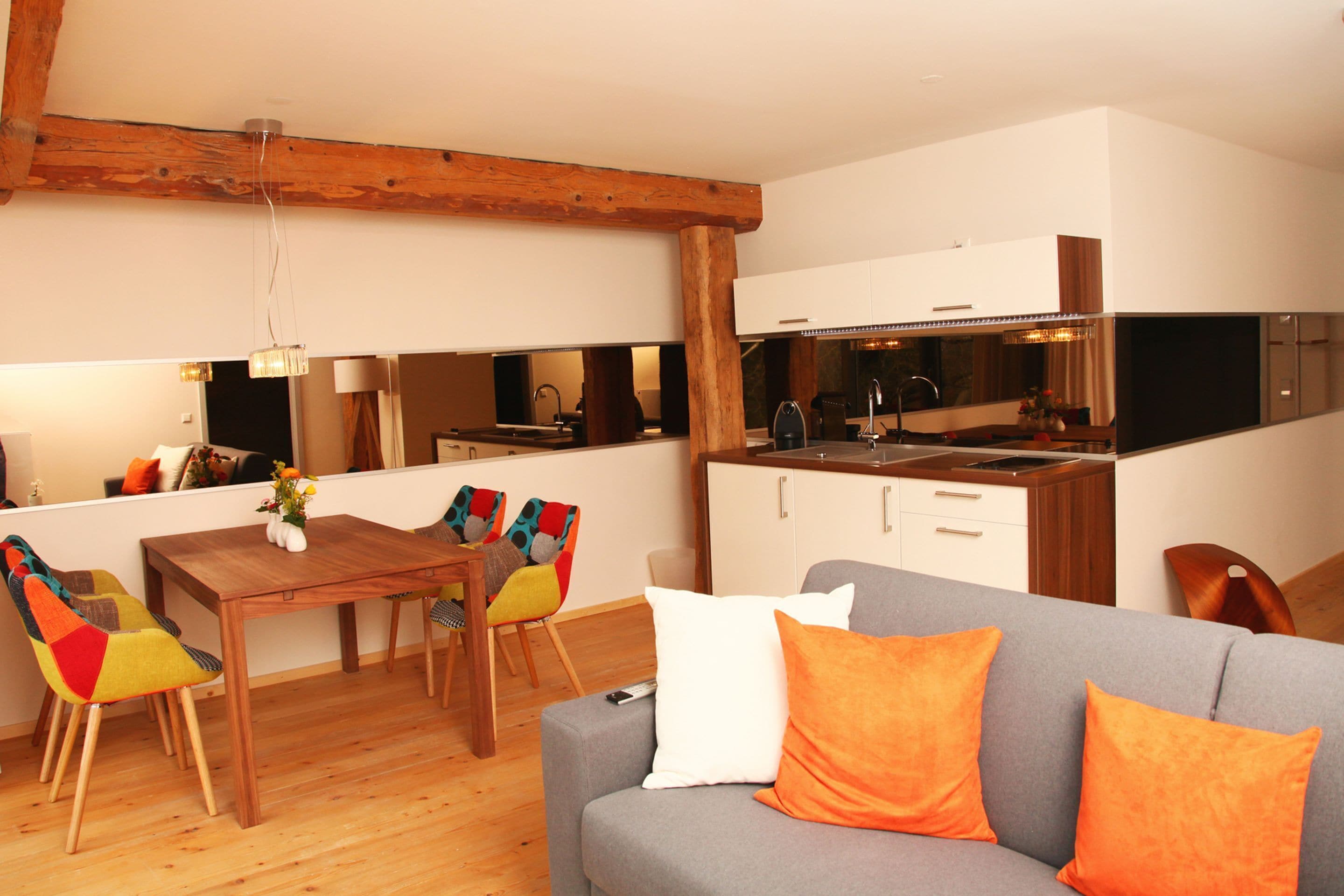 Una sala moderna con un sofá gris, una mesa de comedor de madera, sillas coloridas y una cocina con armarios blancos y techo con vigas de madera.