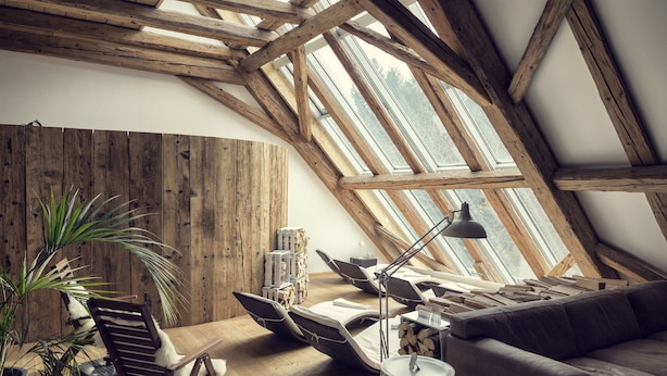 Una habitación estilo loft con techo inclinado, vigas de madera, un tragaluz grande, un sofá, una silla, una lámpara y una planta en maceta.